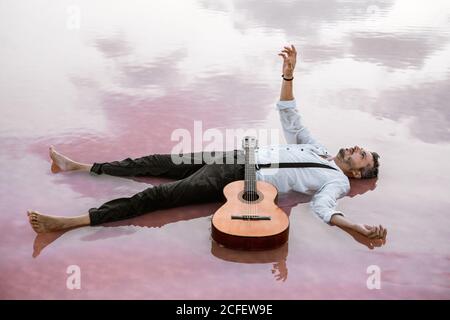 Von oben Mann in weißem Hemd und Strumpfhalter liegen auf Zurück wegschauen und schweben mit akustischer Gitarre und Outstretching Hand an den Himmel im Meer an der Sandbank Stockfoto