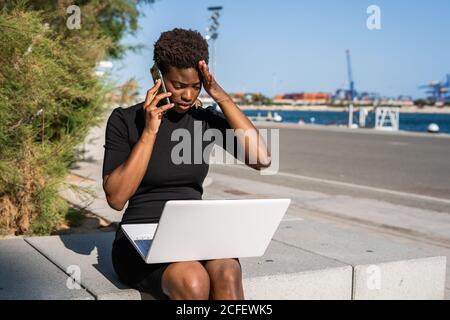 Enttäuschte afroamerikanische Frau in schwarzem Kleid mit Laptop und Gespräch auf dem Smartphone auf der Straße auf verschwommenem Hintergrund Stockfoto