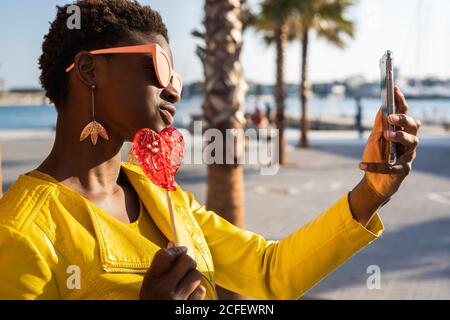 Trendige afroamerikanische Frau in Sonnenbrille in gelber Jacke genießt herzförmigen Lollipop durch Holzzaun Selfie mit Handy zu nehmen Stockfoto