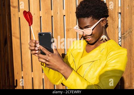 Trendige afroamerikanische Frau in Sonnenbrille in gelber Jacke genießt herzförmigen Lollipop durch Holzzaun Selfie mit Handy zu nehmen Stockfoto