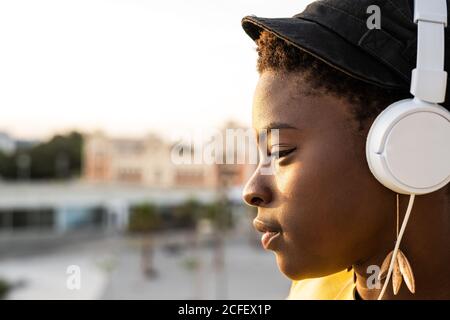 Seitenansicht Inhalt Afrikanisch-amerikanische Frau in modischer Jacke hört Musik in Kopfhörern, während sie sich auf dem Glasbalkon lehnt und auf verschwommenen Hintergrund schaut Stockfoto
