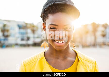 Porträt einer glücklichen afroamerikanischen Frau in stilvoller, heller Jacke, die die Kamera auf dem Sandstrand vor verschwommenem Hintergrund anschaut Stockfoto