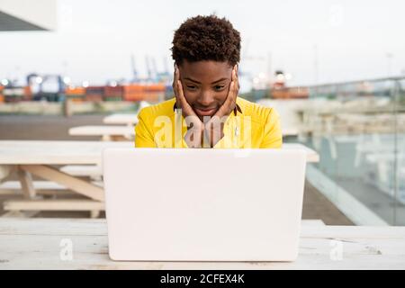 Überraschendes Gesicht einer schwarzen afroamerikanischen Frau in gelber Jacke mit Laptop am Holzschreibtisch in der Stadt auf unscharfem Hintergrund Stockfoto
