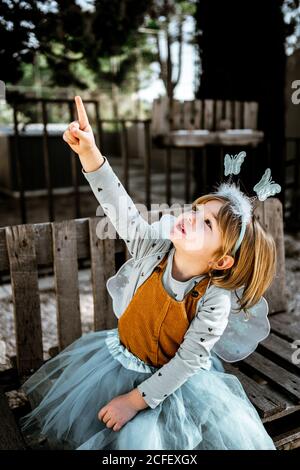 Liebenswert kleines Mädchen in Fee Kostüm sitzt auf schäbigen Holz Bank und Blick auf die Kamera, während Sie Zeit im Hof verbringen Stockfoto