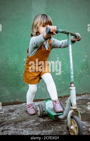 In voller Länge kleine Mädchen in Freizeitkleidung Reiten rostig Kick Roller gegen schäbige grüne Wand auf der Straße Stockfoto