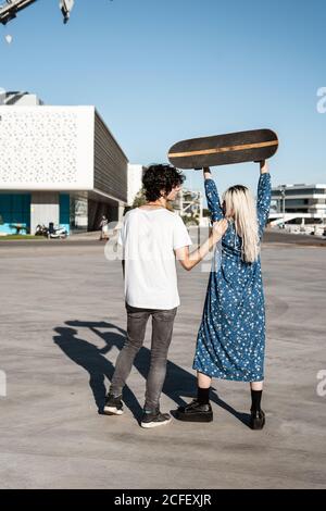 Rückansicht eines nicht erkennbaren jungen trendigen Paares, das steht, während eine Frau ein Skateboard auf dem Platz vor blauem Himmel und verschwommenen modernen Gebäuden bei windigem Wetter hochhebt Stockfoto