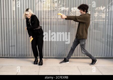 Fröhlicher Mann, der mit Banane auf die Frau zielte, die mit Händen schützte und lachte, während er auf der Straße mit modernem Gebäude im Hintergrund Spaß hatte Stockfoto