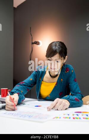 Schöne Frau in elegantem Outfit macht Notizen von Graphen, während Sitzen am Schreibtisch im modernen Büro Stockfoto