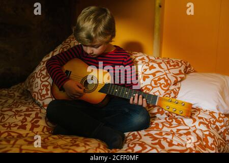 Konzentriertes Kind in lässigem Outfit sitzt auf bequemen Bett in Schlafzimmer und Ukulele spielen, während Sie zu Hause in Kantabrien chillen Stockfoto