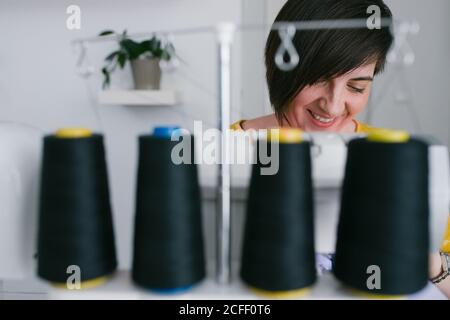 Glückliche Brünette Erwachsene Frau lächeln und mit Nähmaschine, um Kleidung zu machen, während der Arbeit in Hause Werkstatt Stockfoto