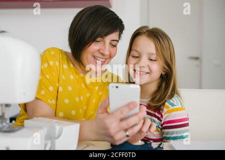 Fröhliche Erwachsene Frau lächelt und nimmt Selfie mit Mädchen, während sie am Tisch sitzt und in der Schneiderei zu Hause arbeitet Stockfoto