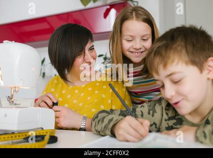 Glückliche Erwachsene Frau und Mädchen lächeln und beobachten Jungen Hausaufgaben machen, während Zeit im Schneideratelier zu Hause zusammen verbringen Stockfoto