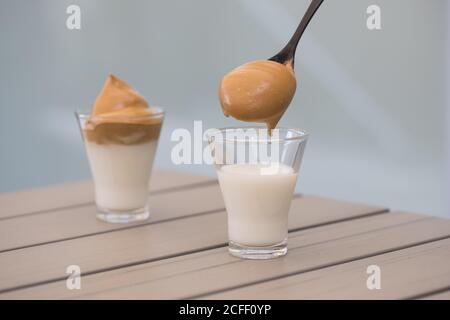 Beschnittene gesichtslose Person Hand mit Teelöffel Zugabe Schlagkaffee in Kalte Milch beim Zubereiten von leckeren Dalgona Kaffee auf Holztisch Stockfoto