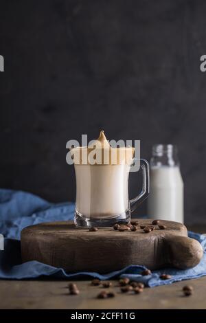 Schlagkaffee Dalgona-Kaffee in kalter Milch in der Nähe von Kaffeebohnen, Teelöffel und Milchflasche auf rustikalen hölzernen runden Bretteroberfläche mit blauer Serviette darunter platziert Stockfoto