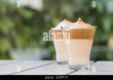 Schlagkaffee Dalgona Kaffee in kalter Milch auf Holztisch Oberfläche mit grünen Pflanzen auf dem Hintergrund Stockfoto