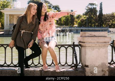 Multiethnische junge Frauen, die lachen und wegschauen, während sie sitzen Geländer und zeigen mit dem Finger in der Nähe des Flusses, während die Zeit zu verbringen Auf dem Damm zusammen Stockfoto