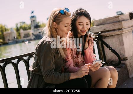 Junge multirassische Frauen in trendigen Outfits lächeln und browsen Smartphone Beim Sitzen in der Nähe von Böschung Geländer an sonnigen Tag auf der Stadt Straße Stockfoto