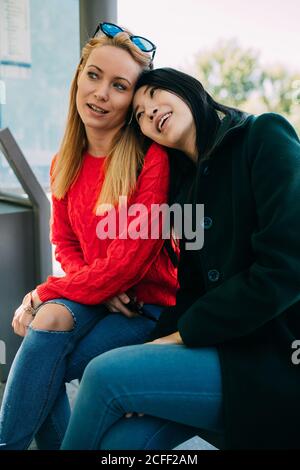 Junge asiatische Frau lächelt und lehnt sich auf die Schulter eines kaukasischen Freundes, während sie auf der Bank sitzt und gemeinsam auf den Bus wartet Stockfoto
