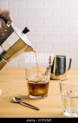 Crop Person Gießen Kaffee aus der Kaffeemaschine in stilvolles Glas mit Eis auf Holztheke im Café Stockfoto