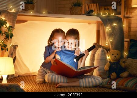 Zwei niedliche kleine Kinder lesen ein Buch mit Taschenlampen im Zelt. Glückliche Mädchen spielen zu Hause. Lustige, nette Kinder, die Spaß im Zimmer haben. Stockfoto