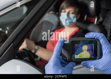 Crop Arzt in medizinischen Handschuhen Messung der Temperatur der weiblichen Fahrer Bei der Verwendung von Infrarotkamera während Coronavirus-Epidemie Stockfoto