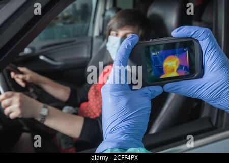 Crop Arzt in medizinischen Handschuhen Messung der Temperatur der weiblichen Fahrer Bei der Verwendung von Infrarotkamera während Coronavirus-Epidemie Stockfoto
