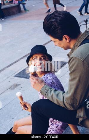 Fröhliche junge attraktive Frau und Freund essen Kornett von Eis, während sie im Freien sitzen Stockfoto