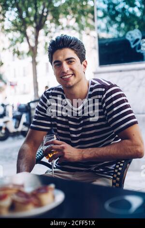 Porträt von fröhlichen jungen Mann in T-Shirt-Stripped Blick auf Kamera, während Sie auf einer Terrasse mit einem Glas sitzen Bier Stockfoto