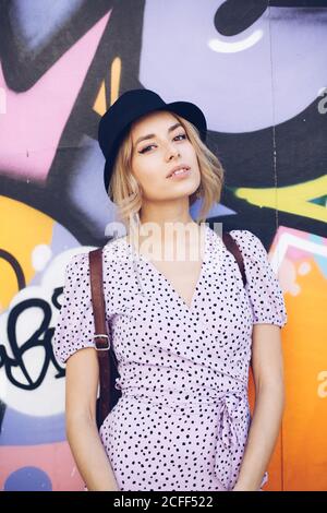 Porträt einer selbstbewussten jungen Frau in Kleid und schwarzem Hut, die auf die Kamera an der Graffiti-Wand blickt Stockfoto