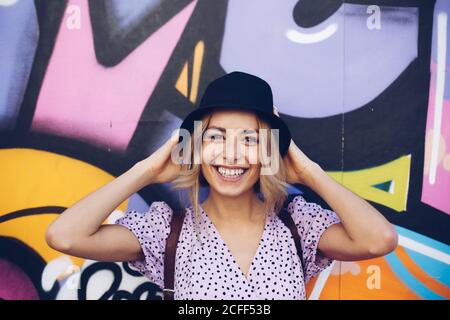 Porträt einer fröhlichen jungen Frau in Kleid und schwarzem Hut, die auf die Kamera an der Graffiti-Wand schaut Stockfoto