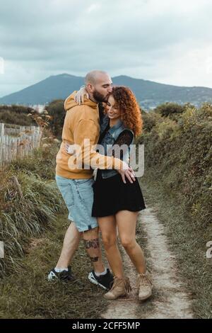 Liebevolle zärtliche Frau berührt das Kinn eines geliebten Mannes, während sie zusammen auf einem Gehweg entlang einer grünen Farm am Meer mit Berg- und Stadtgebäuden im Hintergrund geht Stockfoto