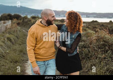 Liebevolle zärtliche Frau berührt das Kinn eines geliebten Mannes, während sie zusammen auf einem Gehweg entlang einer grünen Farm am Meer mit Berg- und Stadtgebäuden im Hintergrund geht Stockfoto