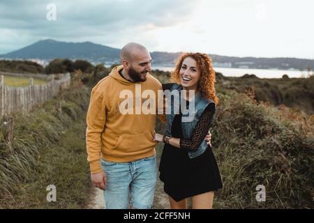 Liebevolle zärtliche Frau berührt das Kinn eines geliebten Mannes, während sie zusammen auf einem Gehweg entlang einer grünen Farm am Meer mit Berg- und Stadtgebäuden im Hintergrund geht Stockfoto