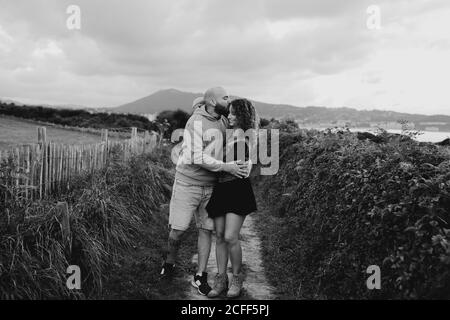 Liebevolle zärtliche Frau berührt das Kinn eines geliebten Mannes, während sie zusammen auf einem Gehweg entlang einer grünen Farm am Meer mit Berg- und Stadtgebäuden im Hintergrund geht Stockfoto