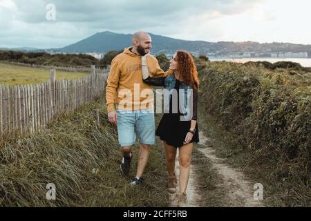 Liebevolle zärtliche Frau berührt das Kinn eines geliebten Mannes, während sie zusammen auf einem Gehweg entlang einer grünen Farm am Meer mit Berg- und Stadtgebäuden im Hintergrund geht Stockfoto