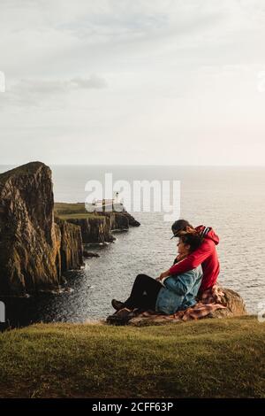 Seitenansicht von jungen Touristen, die auf Karloid sitzen Auf Klippen und umarmen während genießen friedliche Landschaft während Reisen am Scotland Seaside Stockfoto