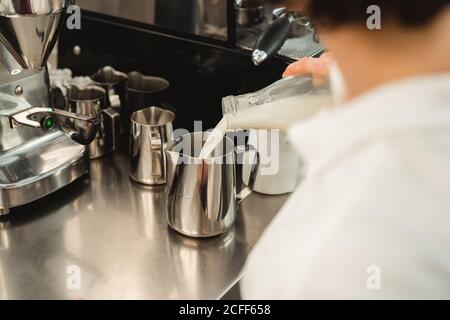 Crop weibliche Barista Gießen Milch aus Glasflasche auf Metall Topf während der Arbeit an der Theke mit Kaffee-Ausrüstung im Café Stockfoto