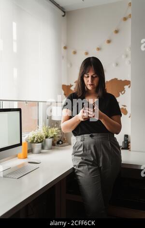 Fokussierte Frau mit dunklen Haaren Messaging auf Handy, während in der Nähe von Desktop-Computer im Büro in Paris Stockfoto