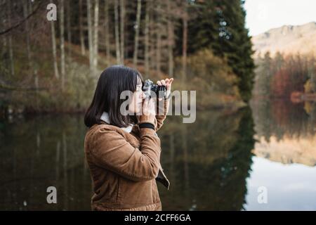 Seitenansicht der jungen weiblichen Reisenden in brauner Jacke nehmen Bild mit Fotokamera beim Stehen neben See umgeben Mit Wald im Herbst Tag Stockfoto