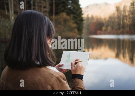 Rückansicht einer Künstlerin, die neben einem schönen See steht Umgeben von Wald im Herbst Tag und Zeichnung in Notizbuch Stockfoto
