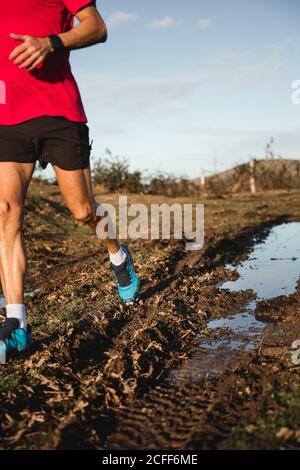 Von unten beschnitten anonyme aktive gesunde Erwachsene männlich in rot shirt und schwarze Shorts Joggen auf Middy Road mit Pfütze Im sonnigen Herbsttag mit blauem bewölktem Himmel im Hintergrund Stockfoto