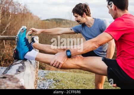 Seitenansicht von müden männlichen Joggern in blau und rot Hemden, die sich nach hartem Laufen und Training am Holzzaun strecken Auf einem grünen Hügel mit schöner Berglandschaft im Hintergrund Stockfoto