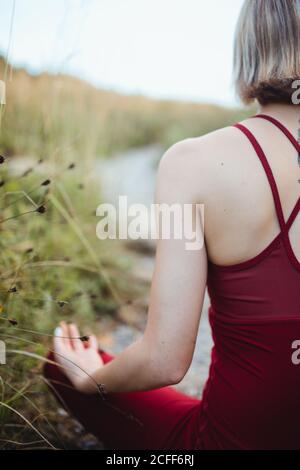 Rückansicht einer nicht erkennbaren Frau in Rot Sportliche Kleidung beim Trainieren und Üben von Lotus Pose Yoga mit Kinn-Mudra-Händen am leeren felsigen Strand Stockfoto