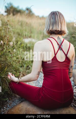 Rückansicht einer nicht erkennbaren Frau in Rot Sportliche Kleidung beim Trainieren und Üben von Lotus Pose Yoga mit Kinn-Mudra-Händen am leeren felsigen Strand Stockfoto
