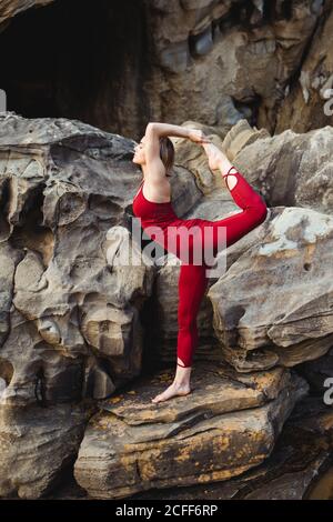 Seitenansicht einer jungen Frau in roter Sportkleidung, die Yoga in der „lord of the Dance“-Pose macht, die auf einem Bein steht, während sie auf rauem Felsen zwischen Bergen verschiedene Beine ausstreckt Stockfoto