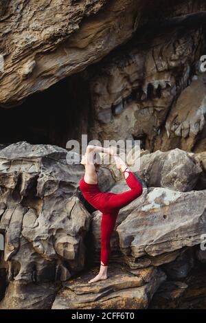 Seitenansicht einer jungen Frau in roter Sportkleidung, die Yoga in der „lord of the Dance“-Pose macht, die auf einem Bein steht, während sie auf rauem Felsen zwischen Bergen verschiedene Beine ausstreckt Stockfoto