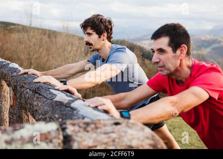 Seitenansicht von müden männlichen Joggern in blau und rot Hemden, die sich nach hartem Laufen und Training am Holzzaun strecken Auf einem grünen Hügel mit schöner Berglandschaft im Hintergrund Stockfoto