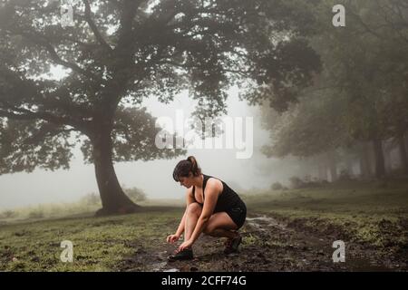 Seitenansicht einer fetten Frau in Sportkleidung, die Schnürsenkel auf Turnschuhen bindet, während sie während des Trainings im nebligen Wald Pause hat Stockfoto