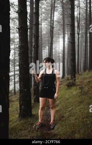 Müde Sportlerin im aktiven Ohr auf engem Pfad im nebligen Wald stehend und mit dem Smartphone beim Liedern genießen und beim Training Pause machen Stockfoto