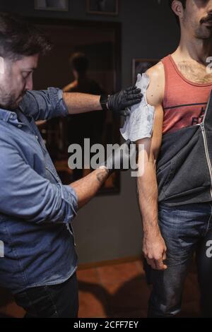 Crop Seitenansicht der konzentrierten männlichen Tätowierer kleben Tattoo Vorlage auf Kundenschulter während der Vorbereitung auf die Arbeit im Salon Stockfoto